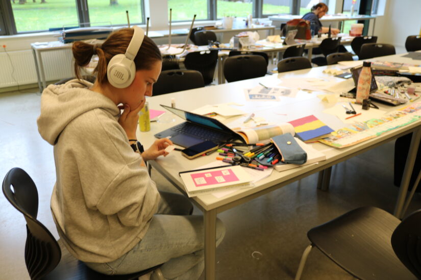 En person sitter ved et bord med laptop, hodetelefoner og mange fargepenner, papirer og tegnesaker spredt utover arbeidsområdet.