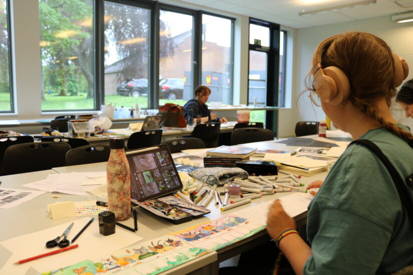 En person sitter med ryggen til foran et arbeidsbord fylt av pc, malerkoster, tegninger, mm.