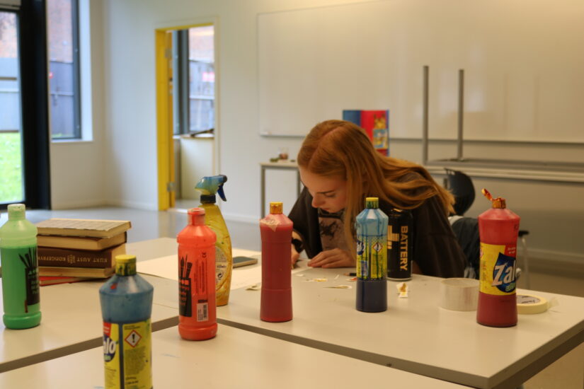 To studenter jobber ved et bord fylt med fargesprutflasker og pensler i et lyst kunstlokale.
