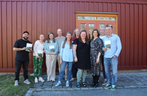 10 mennesker står foran en rød husvegg, noen holder en bok i hånda.