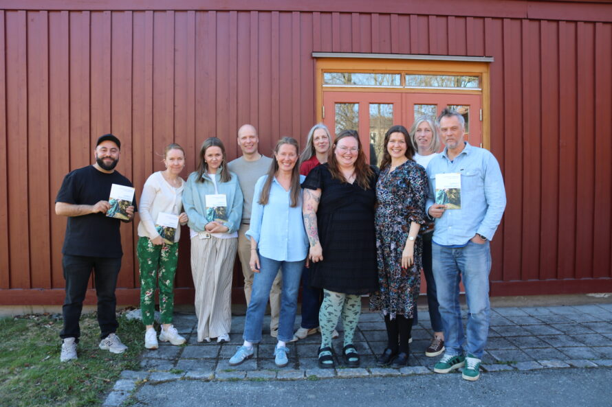 10 mennesker står foran en rød husvegg, noen holder en bok i hånda.