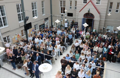 - Trondheim trenger de flinkeste barnehagelærerne, og de utdannes ved DMMH stor gruppe mennesker står tett i en innendørs atrium eller hall, mange holder papir eller programmer, omgitt av lyse bygninger med vinduer og en inngang i bakgrunnen.