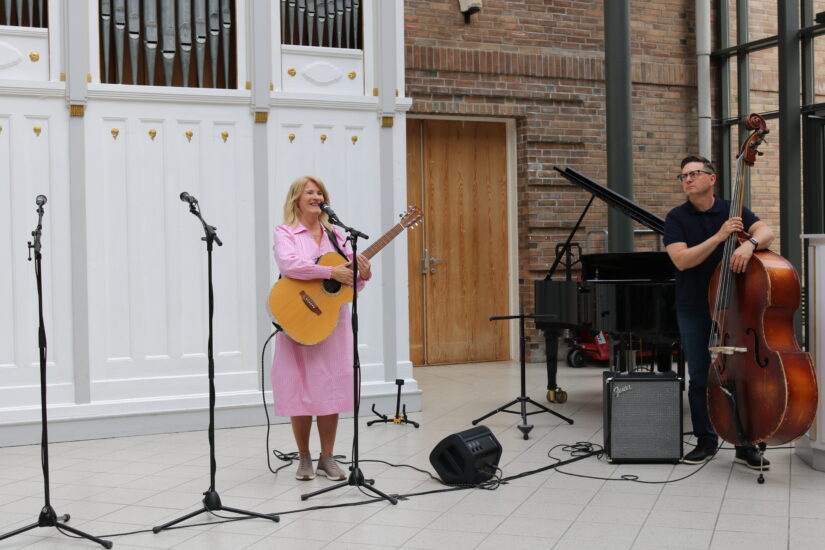 En kvinne i lys rosa kjole spiller gitar og synger inn i mikrofoner, mens en mann spiller kontrabass ved siden av et lite konserthusmiljø med piano og mikrofonstander.