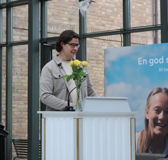 En person står ved en talerstol innendørs, holder en vase med gule roser, mens de ser mot venstre og smiler; bak dem er en blå banner og glassvegger.