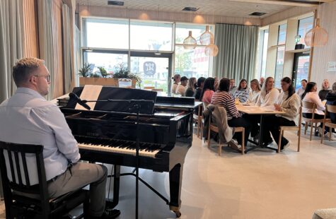 - Dere har valgt en av de viktigste jobbene i samfunnet. Vi ser ryggen til en mann som sitter foran et piano, i bakgrunnen er det to bord som flere folk sitter rundt.