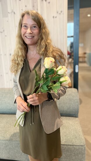 Smilende kvinne med lange krøllete blondt hår bærer en lys blazer og en grønnlig kjole, holder rosa- og hvite roser i armkroken i et innendørs miljø.