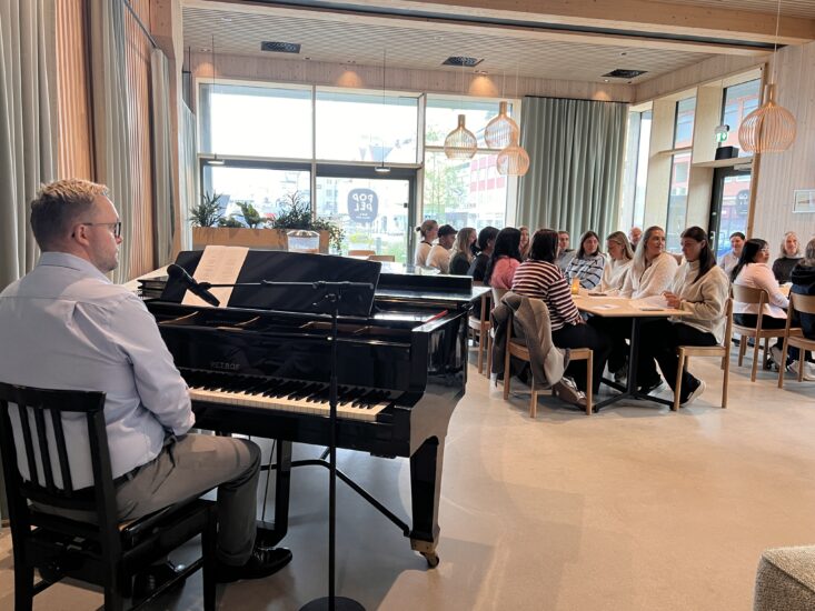 Vi ser ryggen til en mann som sitter foran et piano, i bakgrunnen er det to bord som flere folk sitter rundt.