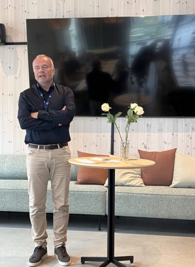 En mann står med armene krysset ved en hørt bord med vase med to lyserosa roser i et rom med lys trepanel og en stor skjerm/TV bak.