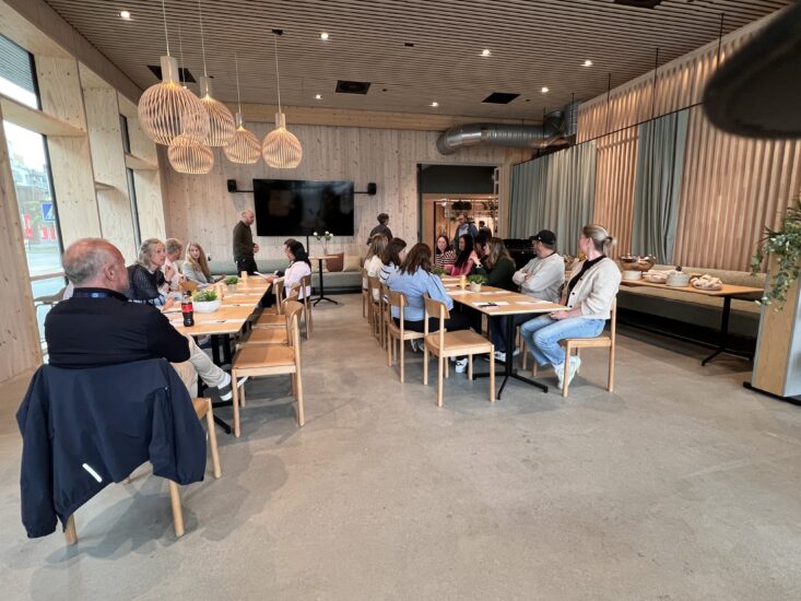 langt bord av mennesker som sitter i konferanse-/møterom, spiser og prater, mens en person står ved et lerret på en kontrastfylt, moderne treetasjersvegg og hengende lamper i taket.