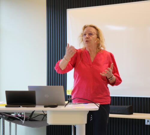 En kvinne står foran en hvit skjerm og prater.