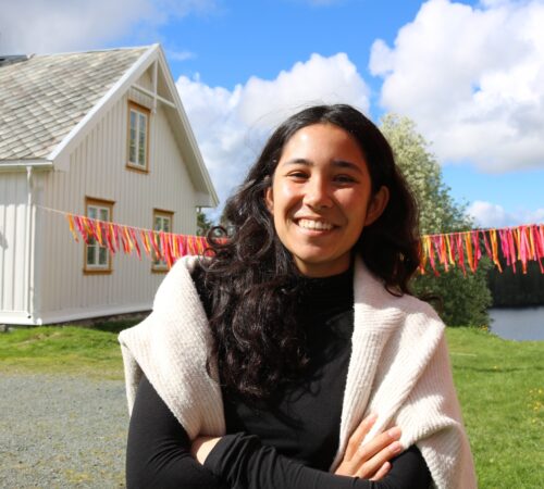 Smilende person står utenfor et lyst hus med fargerike bånd i bakgrunnen, grønt grass og blå himmel med skyer.