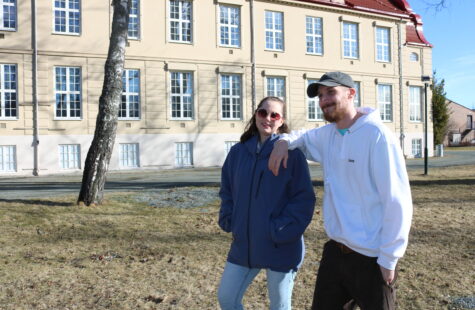 En ung kvinne og en ung mann står ute på en plen foran et stort murbygg i gammel stil