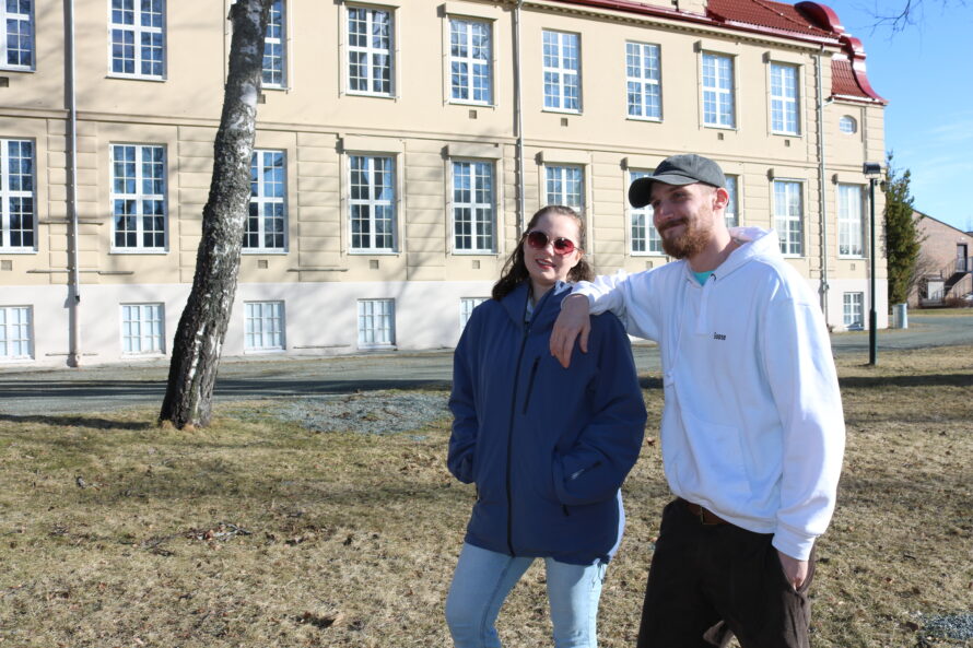 En ung kvinne og en ung mann står ute på en plen foran et stort murbygg i gammel stil