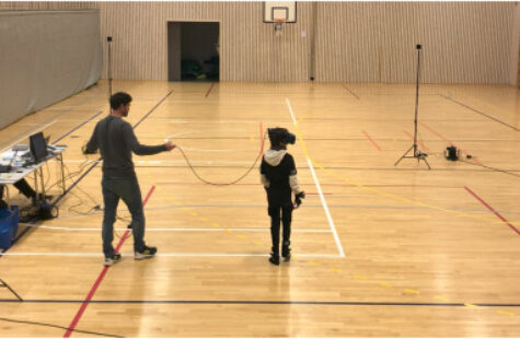 Bilde av en gymsal der et barn står med VR-briller på. Noen voksne sitter langs et bord.