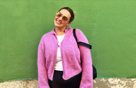 En kvinne i rosa jakke og solbriller står foran en grønn vegg.