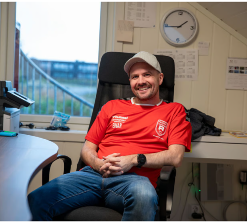 En mann med skyggelue sitter i en kontorstol på et kontor.