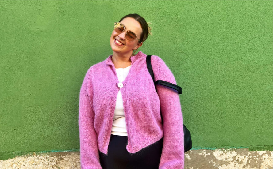 En kvinne i rosa jakke og solbriller står foran en grønn vegg.