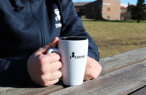 Kopp, hvit med svart innside Bilde av en hvit og sort DMMH kopp som blir holdt av en student på et bord.