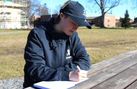 Notatbok Kvinnelig student skriver i DMMH notatbok foran hovedbygget kledd i DMMH merch.