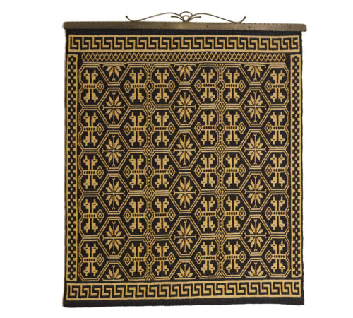 Bilde av vev utført i sort og gul oker med ornamenter. 78 x 91 cm. Plassert ved korridor i mellombygg ved toalett 2. etasje.