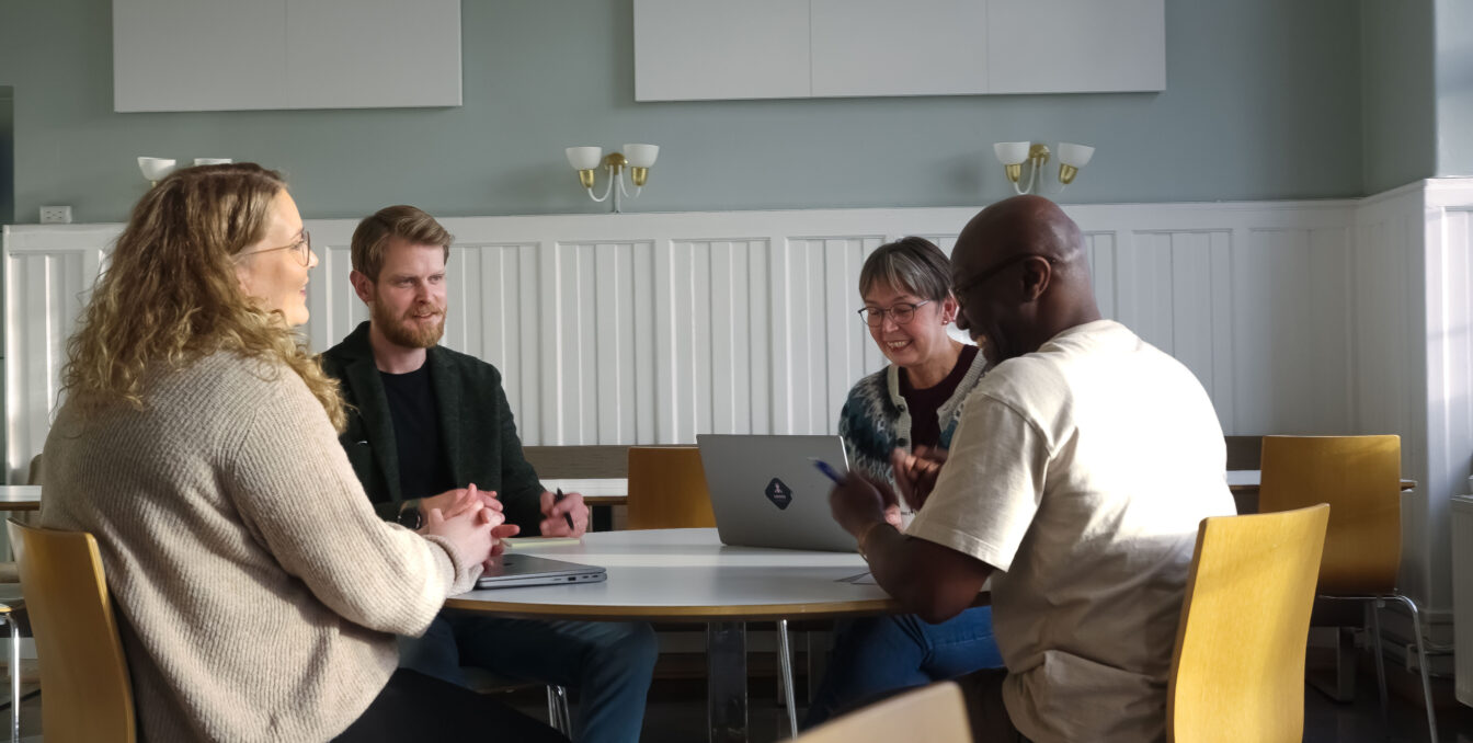 fire personer sitter rundt et rundt bord i et møterom og snakker, med laptop og notatblokk fremme, i et rom med lysegrå vegger og hvite paneler.