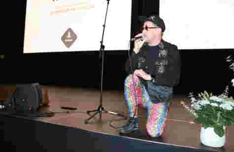 En mann med tights, cap og solbrikker kneler på en scene og synger i mikrofon