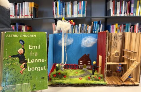 Aåpent bokstativ med en grønn bok av Astrid Lindgren til venstre og en illustrert bokskole med en smålandskap i midten; små figurer i mini-diorama og lekepersoner i treverk til høyre.