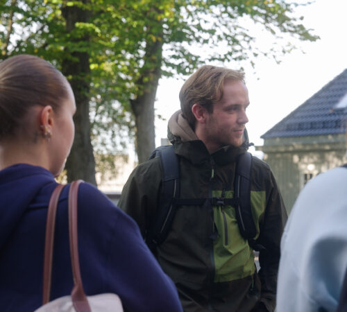 Treffende gruppe ute blant trær, tre personer som snakker, alle med ryggsekker og jakker; sollys filtreres gjennom løvverk.