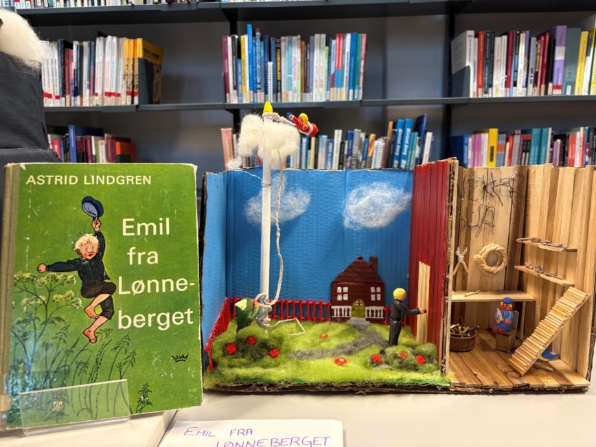 Aåpent bokstativ med en grønn bok av Astrid Lindgren til venstre og en illustrert bokskole med en smålandskap i midten; små figurer i mini-diorama og lekepersoner i treverk til høyre.