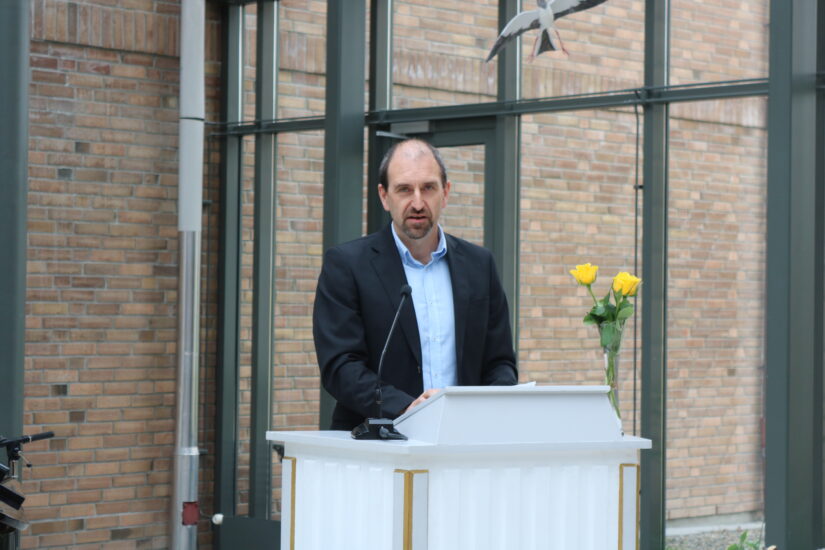 En mann står ved en talerstol, ved siden av tre gule roser.