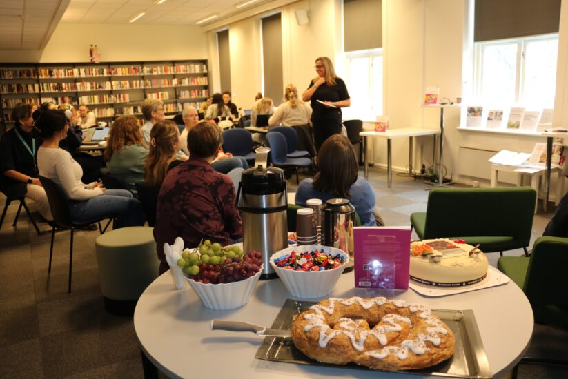 Bilde av et rom med bokhyller og mennesker, i forgrunnen er det et lite bod med kake og frukt på.