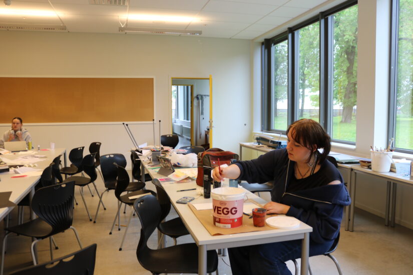 En fargerik kreativt arbeidsplass i et rom med store vinduer, mennesker som maler og tegner ved grupperte bord, papir og maling spredd overalt.