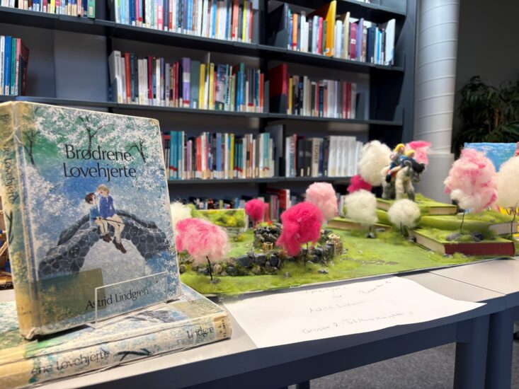 åpne bokutstilling i bibliotek med en stor illustrert bok i forgrunnen og fargerike modeller av trær laget av bomullsull og minidrømmer i grønt landskap på bordet; hyller med bøker i bakgrunnen.