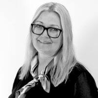 Kvinne med langt lys hår, briller og svarte klær, har et halvt smil; bruker sløyfe rundt halsen og står foran en lys bakgrunn.