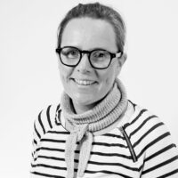 Smilende person med briller, kort/tilbakeført hår, striped svart- og hvit skjorte og en tykk, lysgrå skjerf knytt rundt halsen.