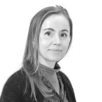 Nærbilde av en person med skulderlangt ligger. Personen har lyst hår, svarer rolig til venstre, iført svart cardigan og mønstret skjorte i loddrett mønster.